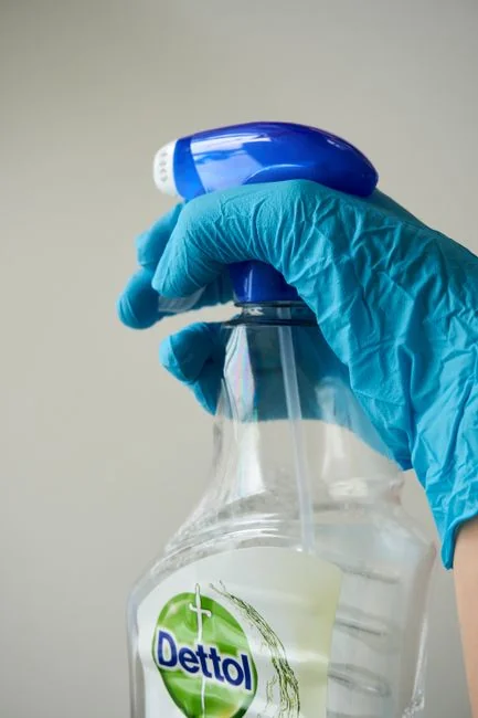 A woman wearing a plaid shirt cleans a kitchen counter using a spray bottle and cloth.